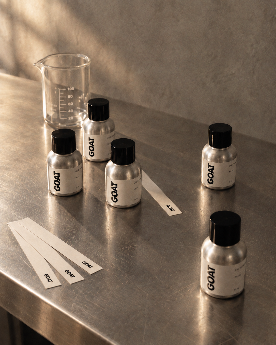 GOAT fragrance oil bottles and scent testing strips on a softly lit stainless steel laboratory bench